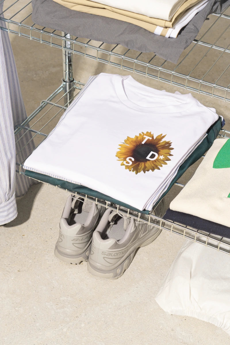 White t-shirt with a yellow sunflower graphic on the chest, on a metal shelf with shoes underneath