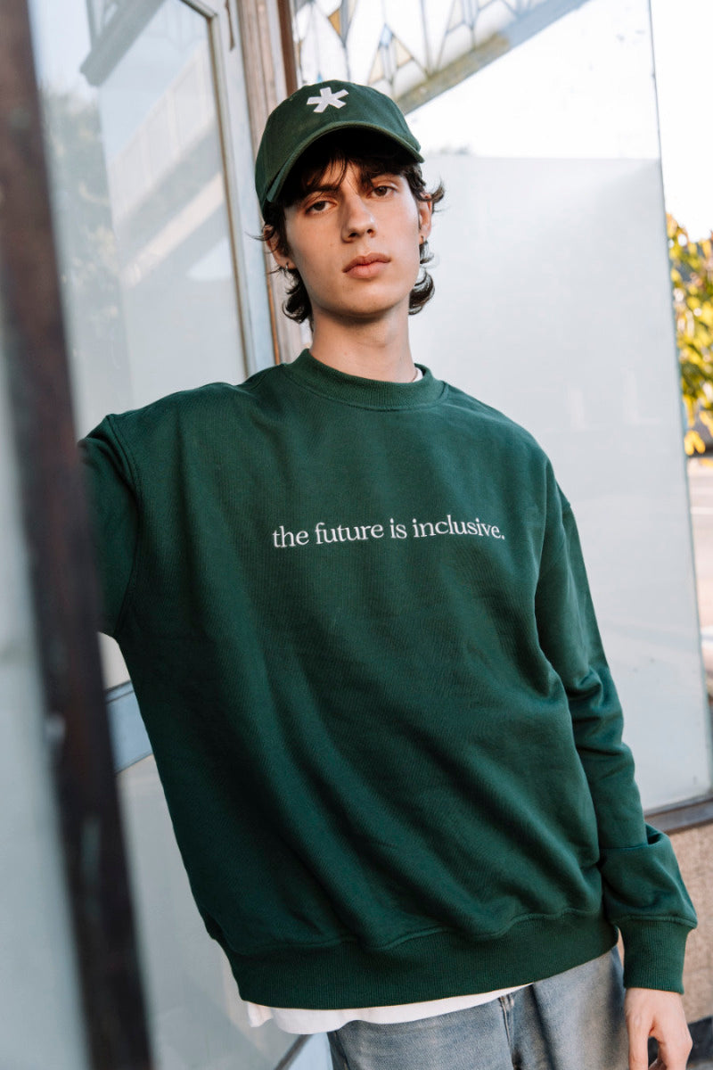 Person wearing a hunter green sweatshirt with 'the future is inclusive' text, standing outdoors.