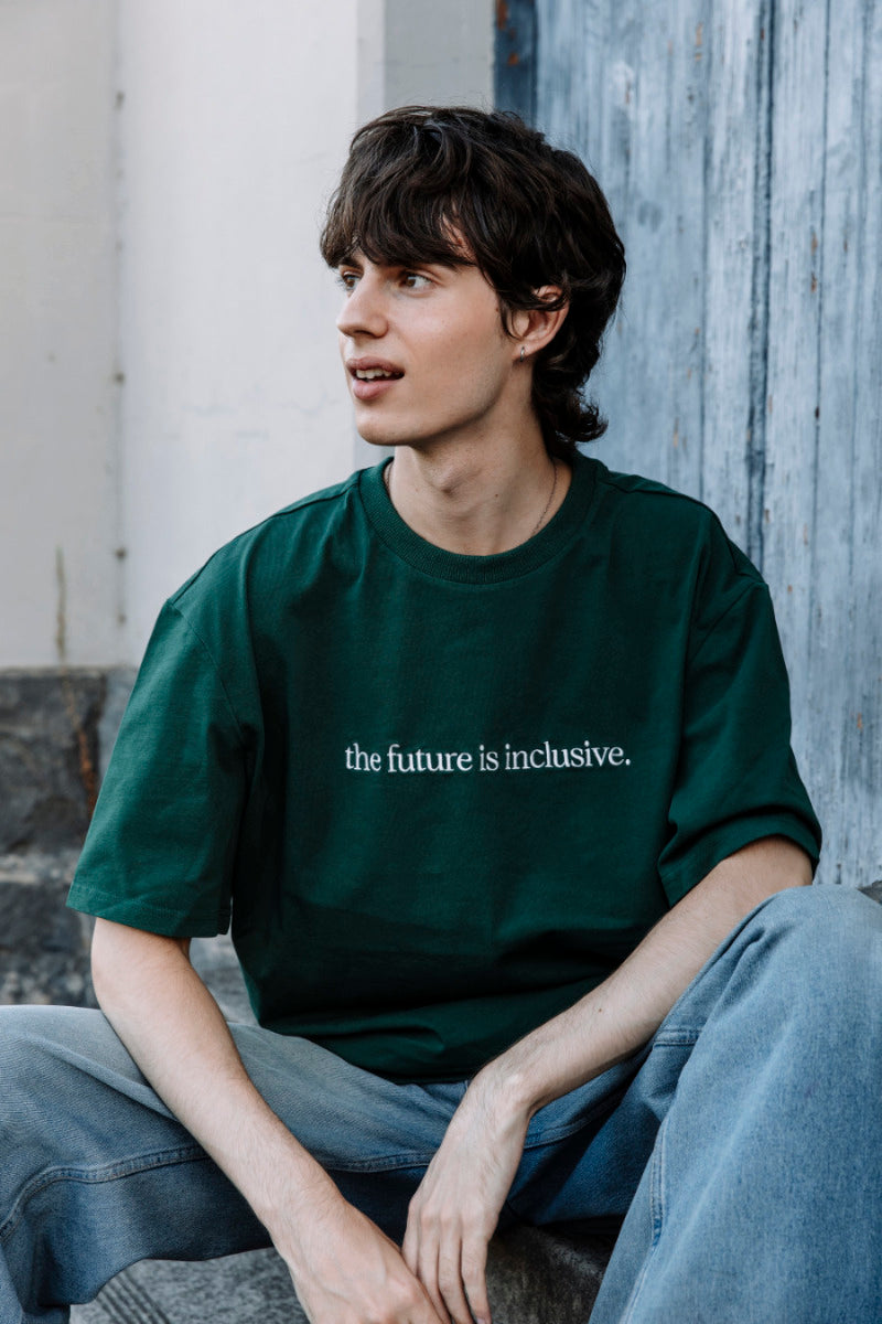 Person wearing a green t-shirt with 'the future is inclusive.' text, sitting against a textured wall.