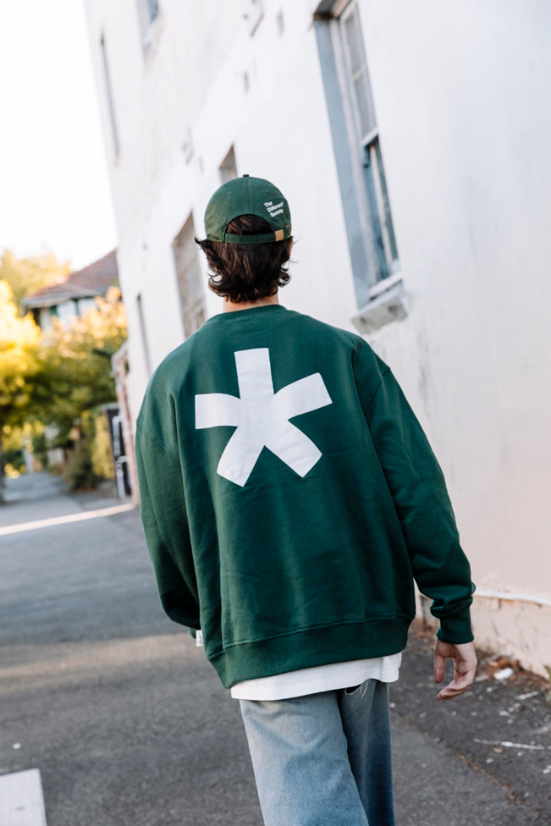 Person wearing a green sweatshirt with a white asterisk logo on the back, walking on a street.