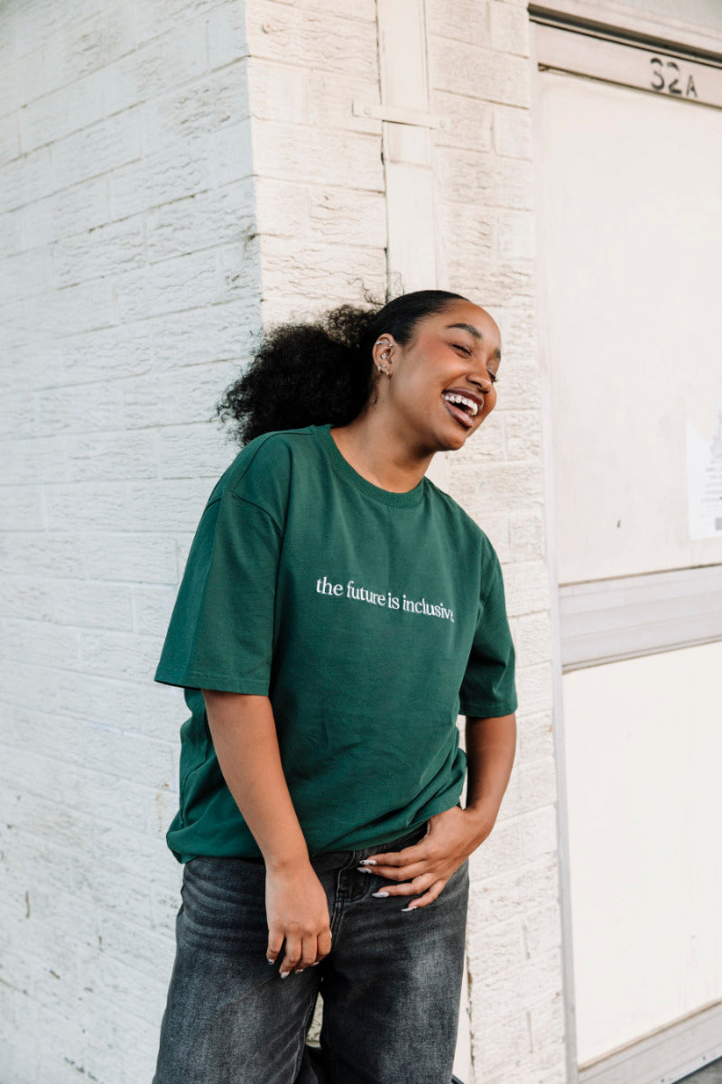 Person wearing a green t-shirt with the text "the future is inclusive", standing against a white brick wall.