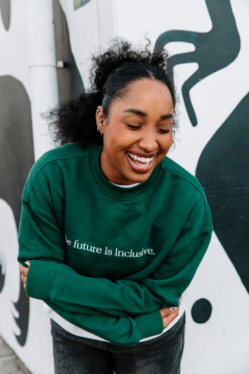 Person wearing a hunter green sweatshirt with 'The future is inclusive' text, standing against a wall with abstract designs.