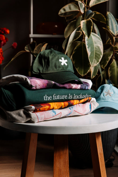 Stack of The Different Society sweatshirts, t-shirts and caps on a table, with plants in the background
