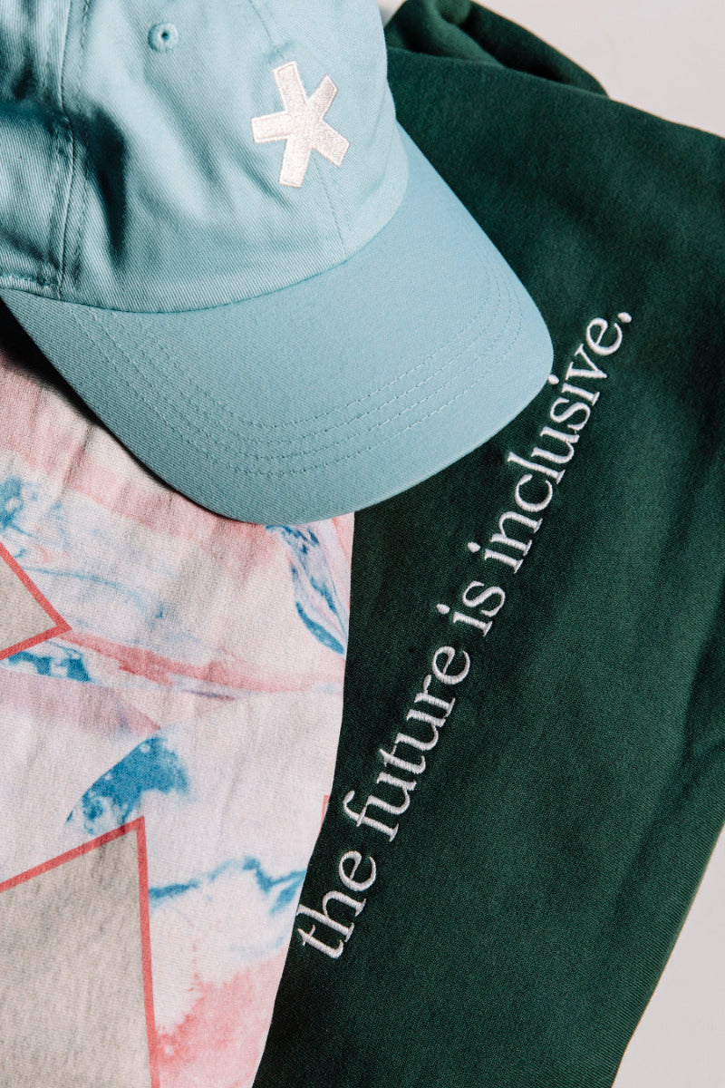 Light green dad cap featuring The Different Society's "asterisk" logo, sitting on top of a printed t-shirt, and dark green sweatshirt with 'the future is inclusive' text on it