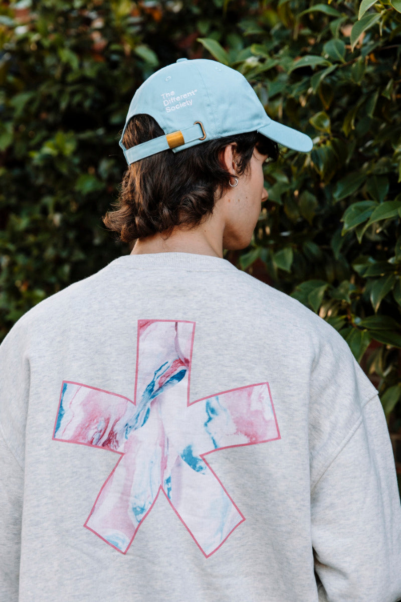 Person wearing a light green cap and gray sweatshirt with a colorful asterisk  logo, standing against a green leafy background.