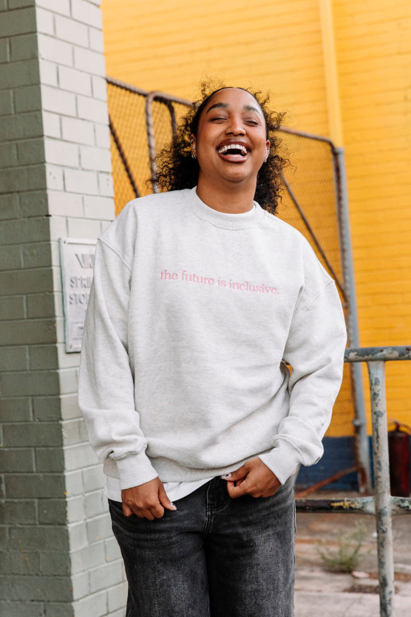 Person wearing a light grey marle sweatshirt with pink text "the future is inclusive", standing in front of a yellow wall.