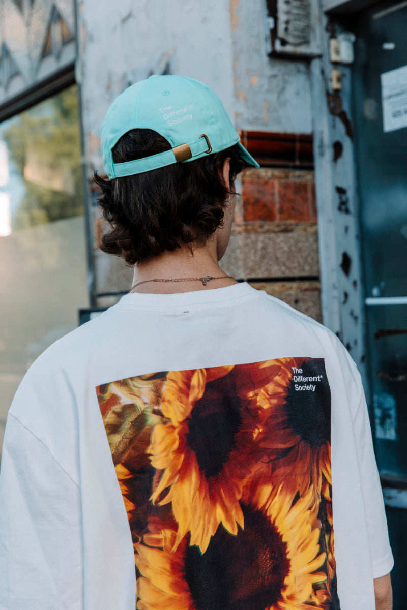 Person wearing a white t-shirt with a sunflower design and a light green cap, standing in an urban setting.