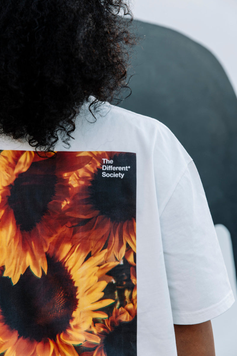 Person wearing a white t-shirt with yellow sunflower design and 'The Different Society' text.