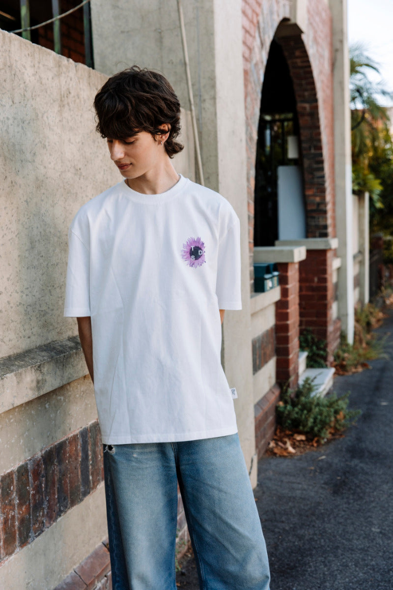 Person wearing a white t-shirt with a purple sunflower design on the left side of the chest, standing outdoors.