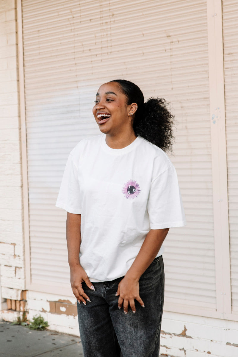 Person wearing a white t-shirt with a purple flower design, standing in front of a white wall.