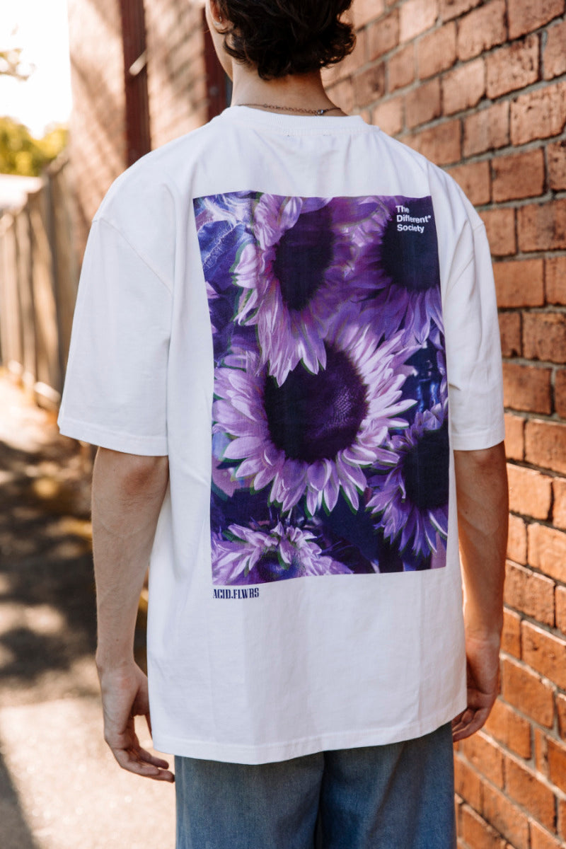 Person wearing a white t-shirt with a purple floral design on the back, standing against a brick wall.