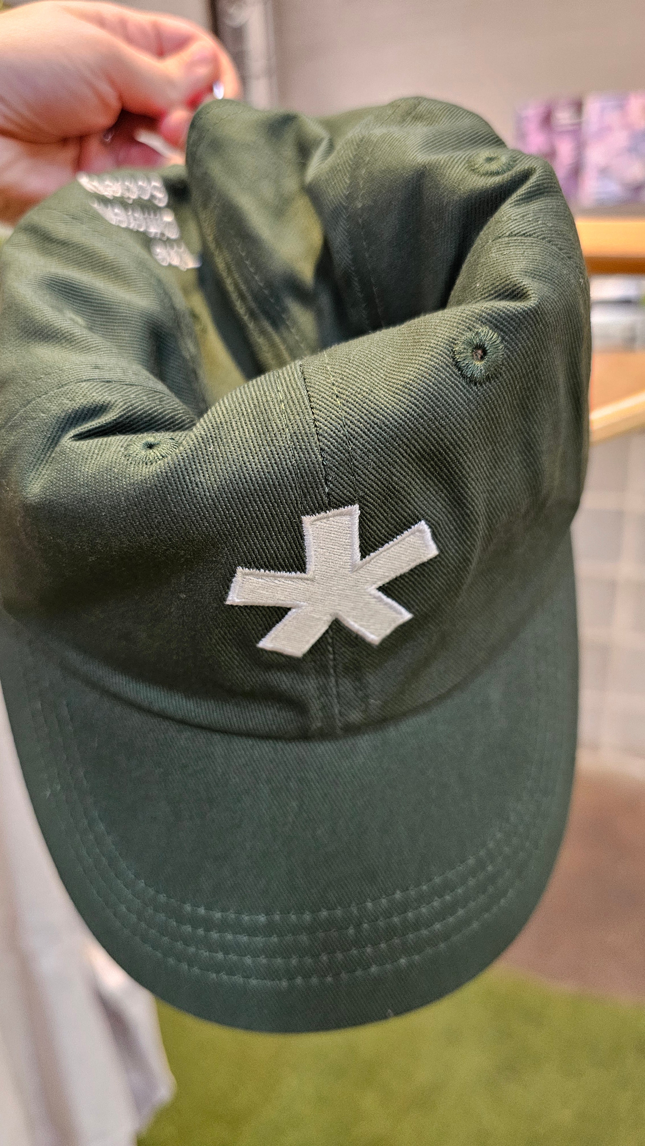 Hunter green cap with The Different Society asterisk logo stitched on the front, held by a hand against a blurred indoor background